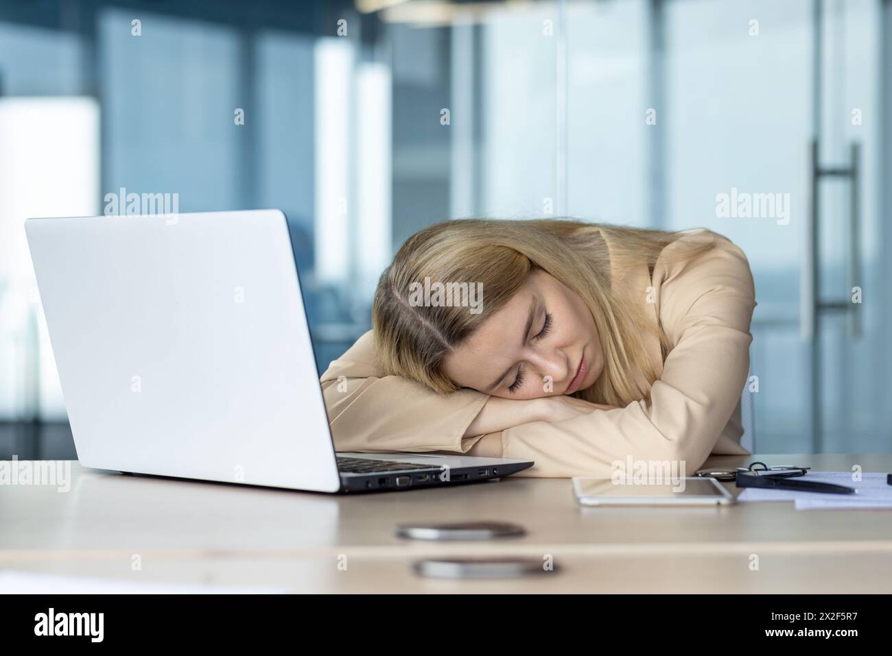 La giovane donna bionda si è trovata in un momento di stanchezza, dormendo alla scrivania circondata da oggetti di lavoro, tra cui smartphone, penne e un laptop. Foto Stock