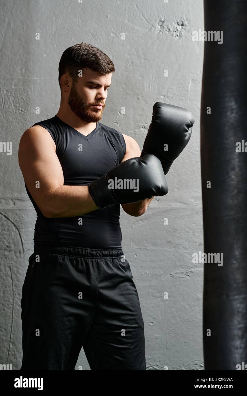 Un bell'uomo con la barba si trova accanto a una borsa da pugno in palestra, praticando tecniche di pugilato con concentrazione e determinazione. Foto Stock