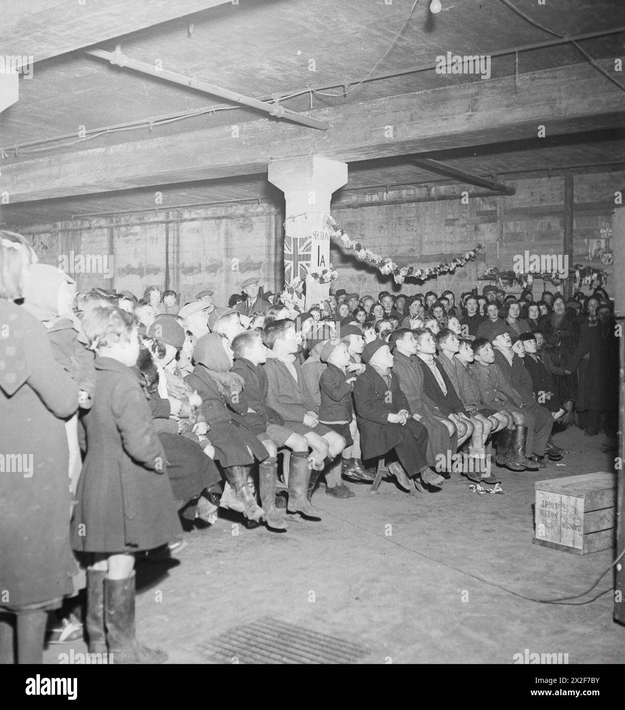 LA PRIMA MOSTRA CINEMATOGRAFICA IN UN CINEMA AIR RAID SHELTER, BIRMINGHAM, INGHILTERRA, 1940 - la fotografia mostra il pubblico, composto in gran parte da bambini, in un grande rifugio appositamente costruito. Sono seduti intorno a una colonna contrassegnata dalla "sezione 1A" e decorata con un Union Jack e un bunding. Né il proiettore né lo schermo sono visibili Foto Stock