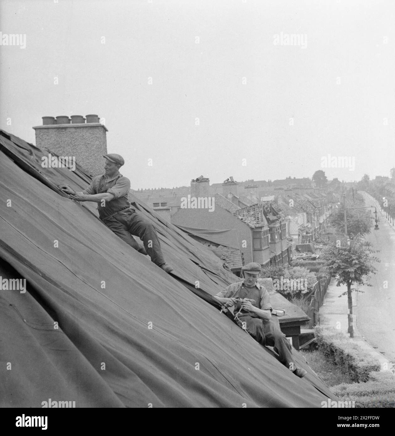 CAMPO DI RIPARAZIONE BLITZ A LONDRA: LA VITA DI TUTTI I GIORNI CON LE SQUADRE DI RIPARAZIONE BLITZ, LONDRA, INGHILTERRA, Regno Unito, 1944 - i costruttori della "Blitz Repair Squad" posero teloni sui tetti delle case per proteggere gli interni dagli elementi a seguito di un attacco V1, che danneggiò la maggior parte delle case della strada. Questi costruttori fanno parte di una grande squadra che si è recata a Londra da varie parti del paese per aiutare a riparare tali danni Foto Stock
