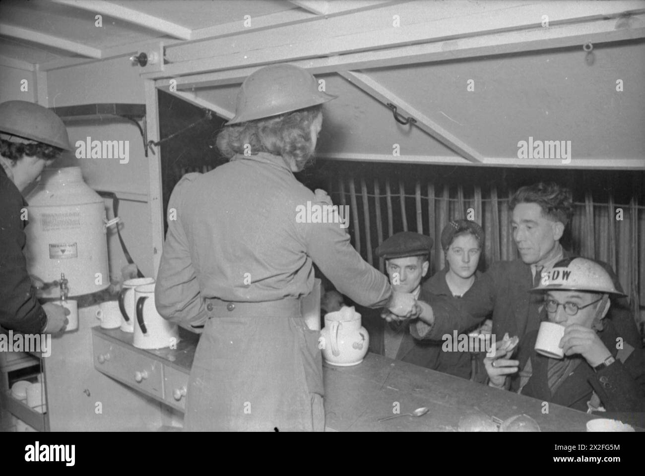 MENSA BLITZ: LE DONNE DEL SERVIZIO VOLONTARIO FEMMINILE GESTISCONO Una MENSA MOBILE A LONDRA, INGHILTERRA, 1941 - Patience 'Boo' Brand serve il tè a un rifugio della mensa WVS, mentre un guardiano ARP beve il suo tè, un panino in mano, sotto la tenda della mensa. Anche Rachel Bingham sta lavorando dietro il bancone, versando il tè da un'urna in una tazza. Tutti i rinfreschi vengono venduti agli scaffali al prezzo di costo e il WVS non produce profitti Foto Stock