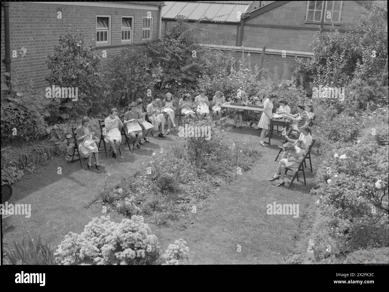 Una FOTO DI Una CITTÀ DEL SUD: LIFE IN WARTIME READING, BERKSHIRE, INGHILTERRA, Regno Unito, 1945 - le ragazze prendono parte a una lezione di maglieria all'aperto al sole nel giardino sommerso della Caversham Modern Secondary School. Ci sono anche due ragazze al lavoro sulle macchine da cucire, sedute ai tavoli che sono stati allestiti sull'erba. Secondo la didascalia originale, "la tendenza è quella di dividere ragazzi e ragazze di undici anni in scuole separate o gruppi separati nella stessa scuola" Foto Stock