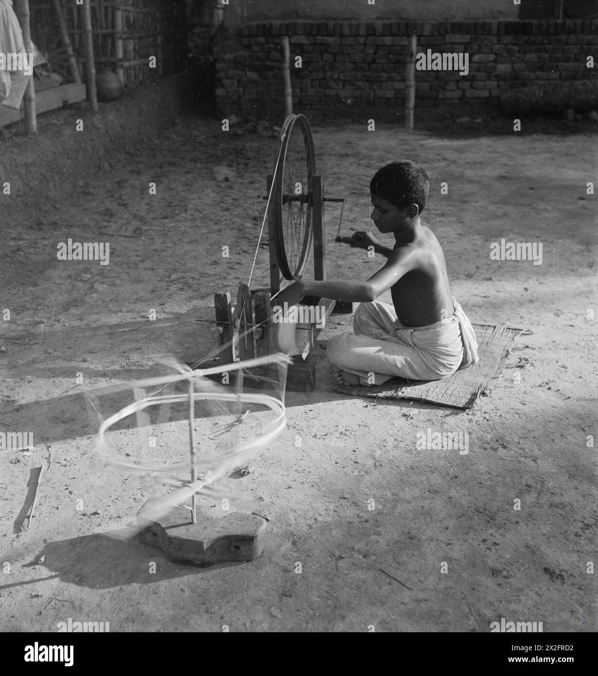 Una fotografia di Cecil Beaton in India, 1944, mostra un ragazzo bengalese seduto su una ruota rotante con il suo filo illuminato dalla luce del sole, catturando la vita quotidiana e le pratiche tradizionali. Foto Stock Una fotografia di Cecil Beaton in India, 1944, mostra un ragazzo bengalese seduto su una ruota rotante con il suo filo illuminato dalla luce del sole, catturando la vita quotidiana e le pratiche tradizionali. Foto Stock