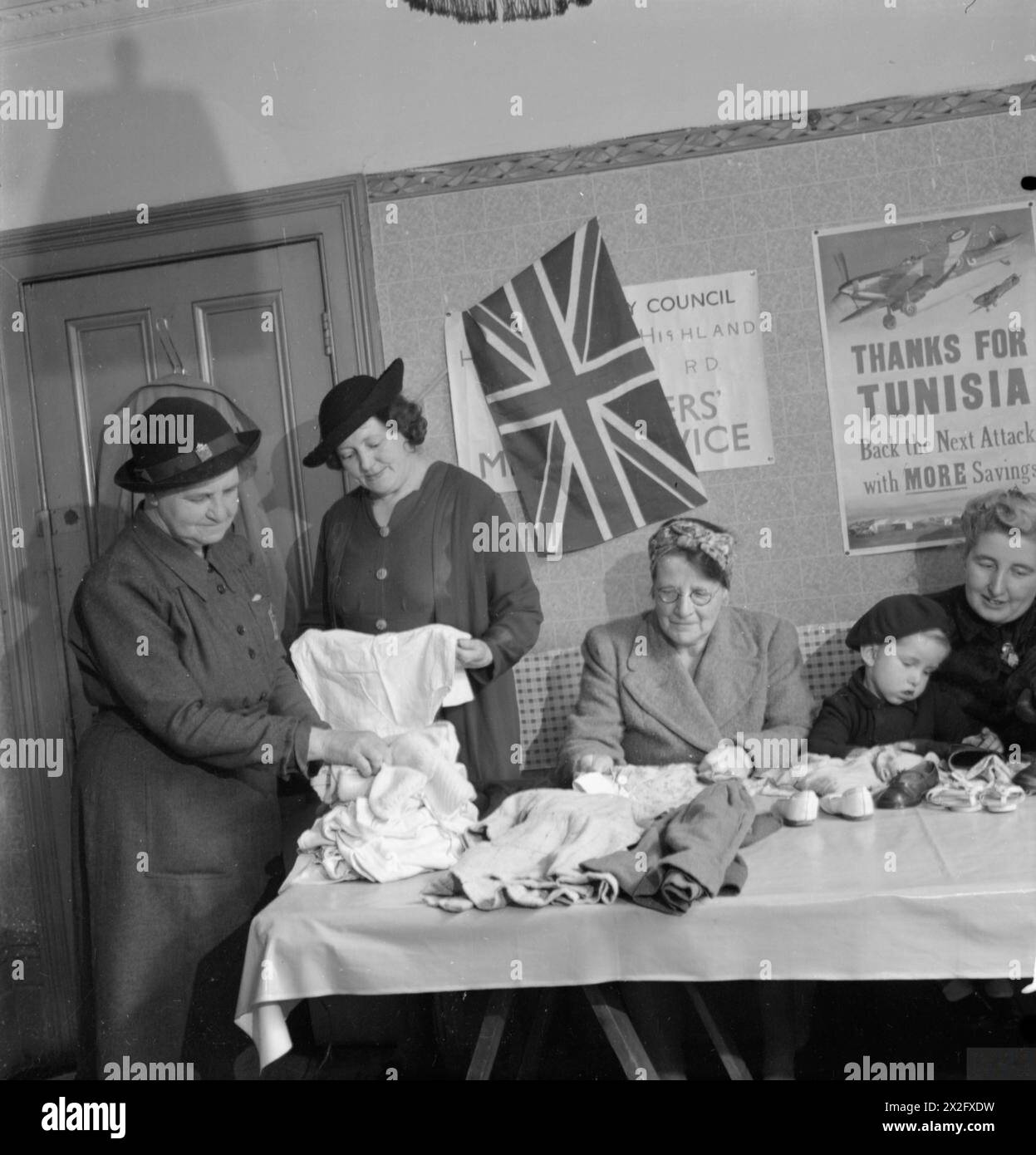Fotografia che mostra un cambio di abbigliamento per bambini a Norwood, Inghilterra, 1943, con i membri del servizio volontario femminile che esaminano i ponticelli sotto un sistema di baratto, decorati con bandiere sindacali e poster. Foto Stock