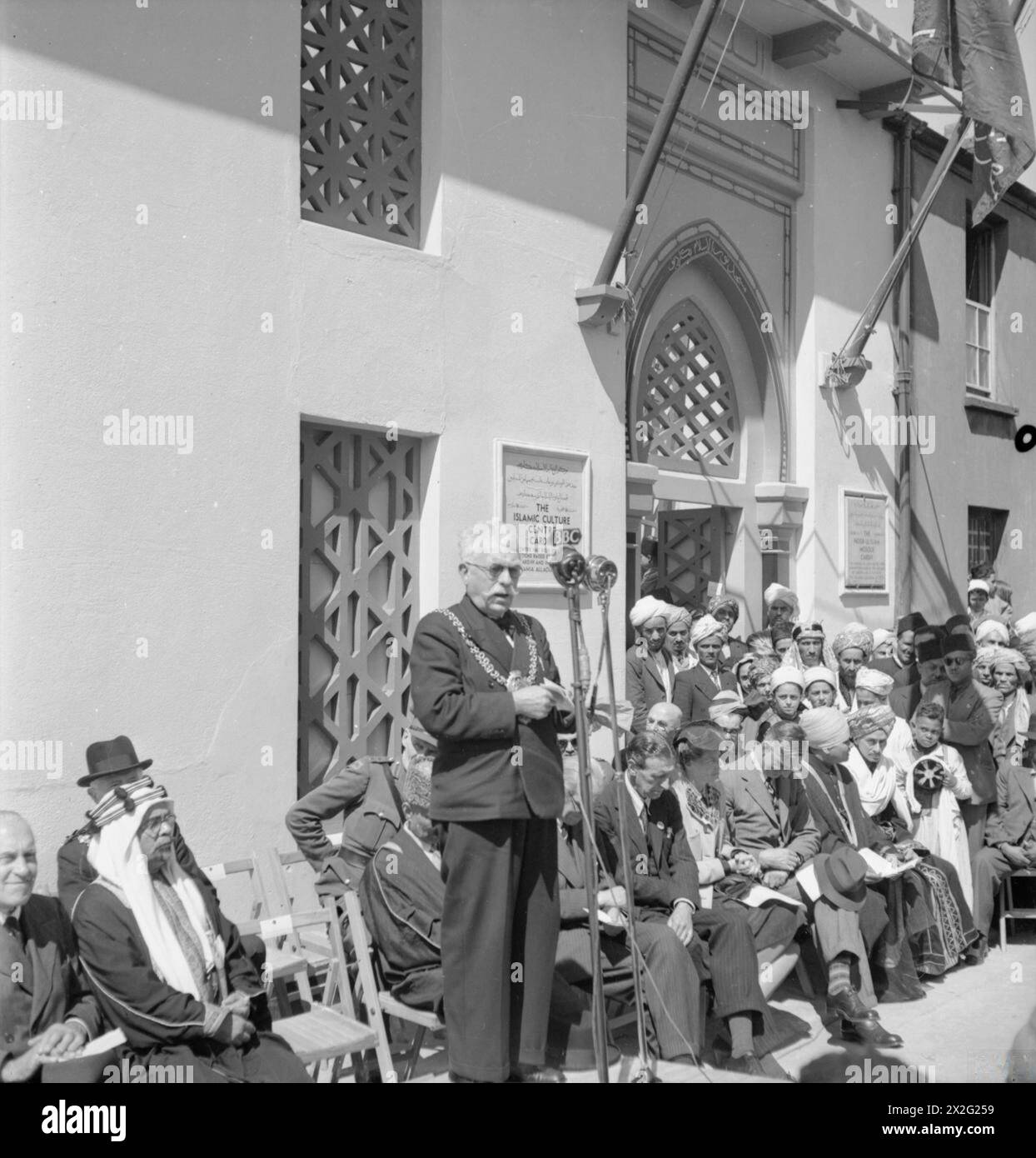 COMUNITÀ MUSULMANA: VITA QUOTIDIANA A BUTETOWN, CARDIFF, GALLES, Regno Unito, 1943 - il sindaco di Cardiff, consigliere James Griffiths, si rivolge al pubblico riunito al sole fuori della nuova Moschea di Peel Street durante la cerimonia di apertura civile. Vari dignitari possono essere visti riuniti accanto a lui intorno all'ingresso principale della moschea, tra cui sua Eccellenza Sheikh Hafiz Wahba, ministro dell'Arabia Saudita. Due targhe, una su entrambi i lati della porta, affermano che la Moschea Noor UL Islam e il Centro culturale Islamico associato sono stati eretti con iscrizioni erette dai musulmani a Cardif Foto Stock