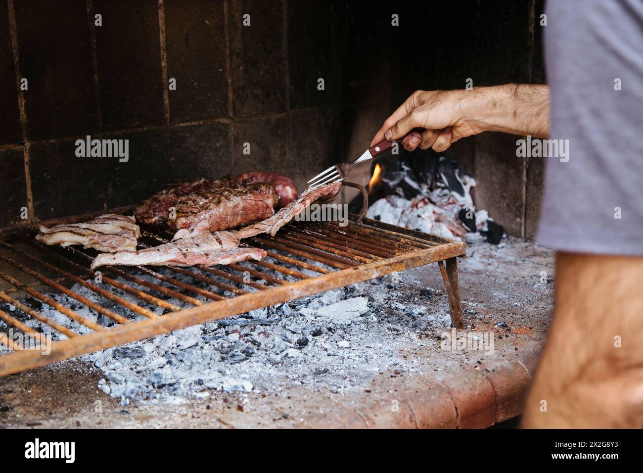 Uomo che cucina la carne sul fuoco, che prepara il barbecue sui carboni nella griglia di casa sua. Asado tradizionale di Argentina, Cile, Paraguay, Uruguay Foto Stock