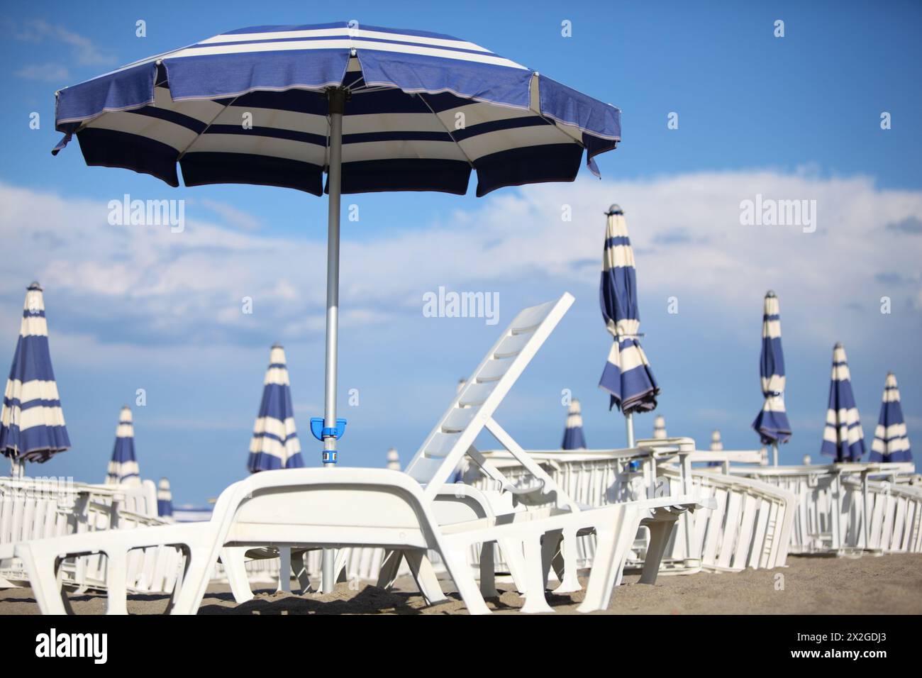 molti lettini bianchi e ombrelloni blu sulla sabbia della spiaggia. cielo blu e nuvole. profondità di campo ridotta Foto Stock