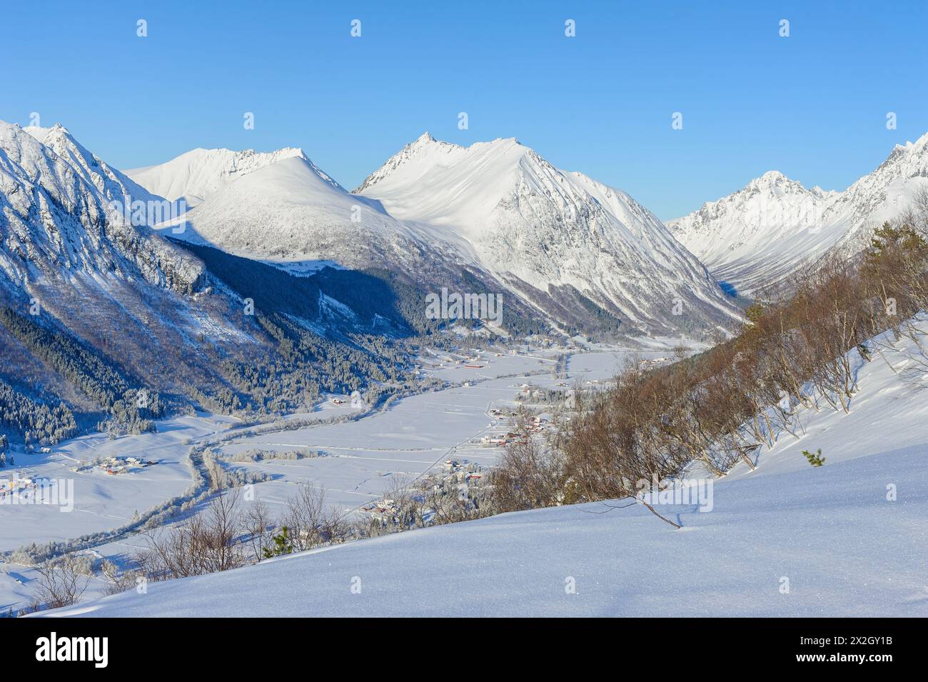 Una vista panoramica di un villaggio annidato in una valle con montagne innevate sotto un cielo azzurro. Foto Stock