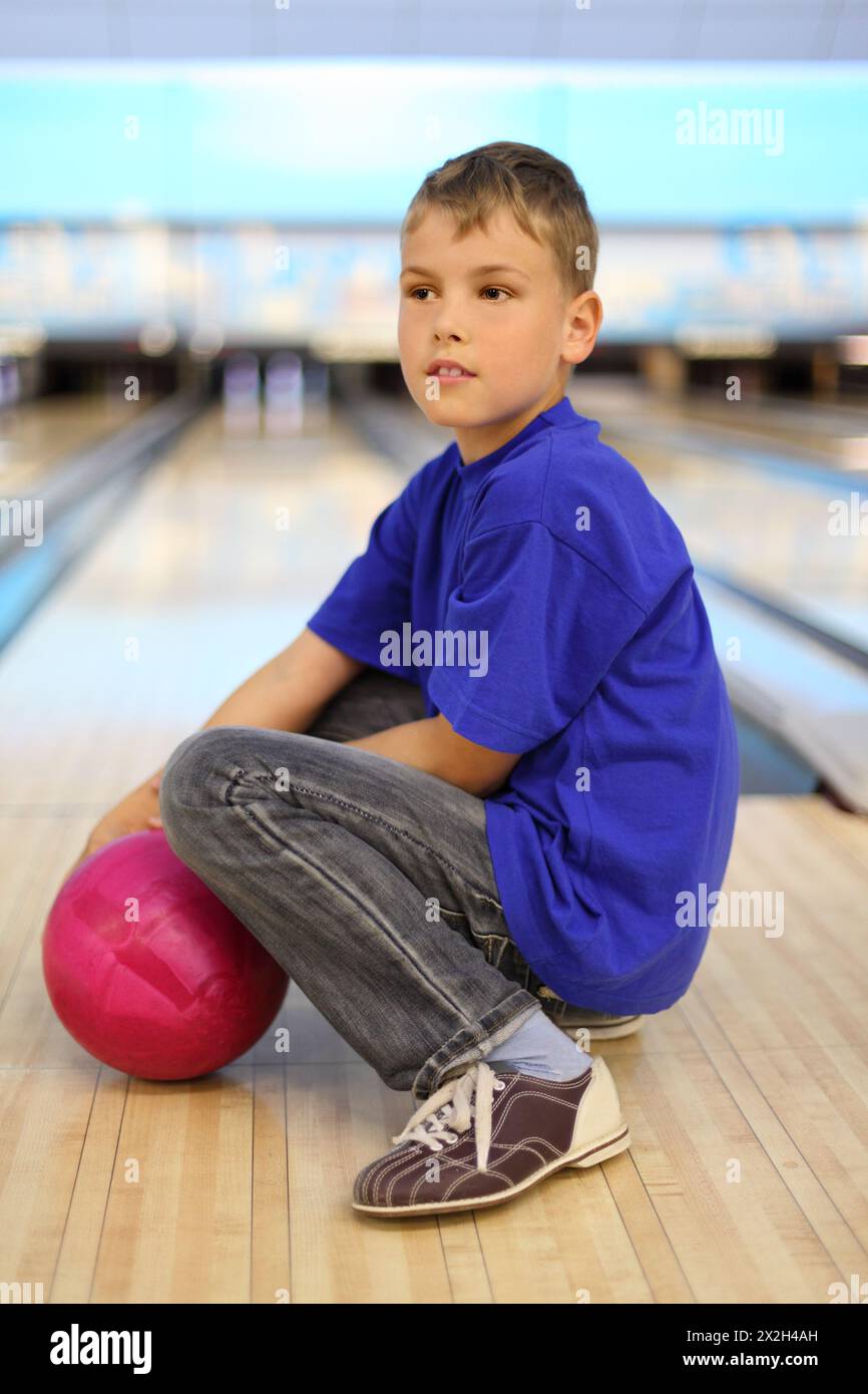 Il ragazzo vestito con una maglietta blu con palla rosa si siede sul pavimento nel club da bowling; profondità di campo bassa; corpo intero Foto Stock