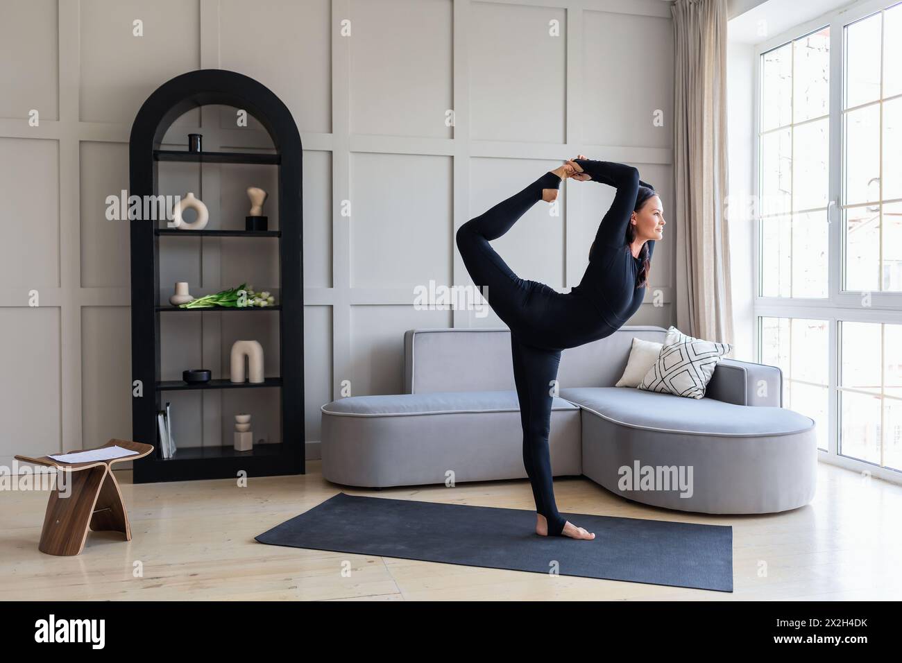 Donna di yoga che esegue una variazione dell'esercizio Natarajasana, re della posa di danza, allenamento in abbigliamento sportivo nero in camera in piedi su un tappetino, lengt pieno Foto Stock