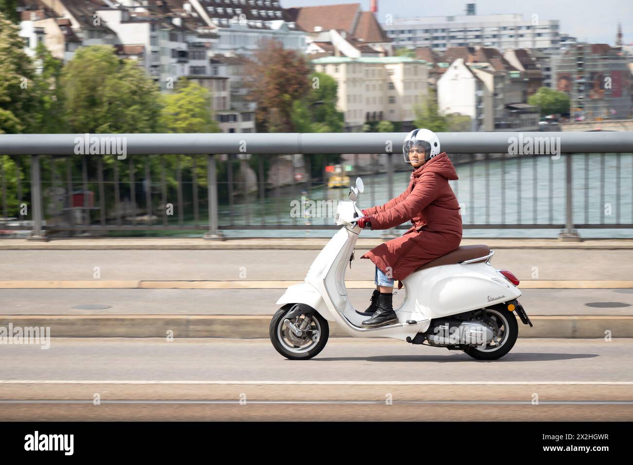 Basilea, Svizzera - 18 aprile 2024: Una donna che indossa un parka in sella a uno scooter bianco su Wettsteinbrucke Foto Stock