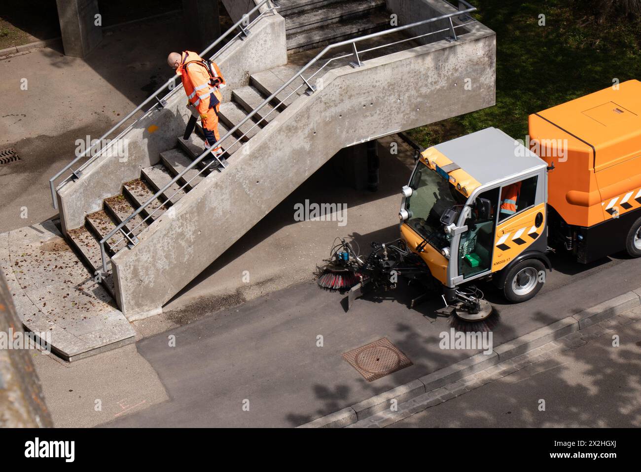 Basilea, Svizzera - 18 aprile 2024: Lavoratore che indossa un'uniforme arancione per la pulizia del ponte cittadino scale con soffiatore di foglie e spazzatrici stradali per la pulizia dei veicoli Foto Stock