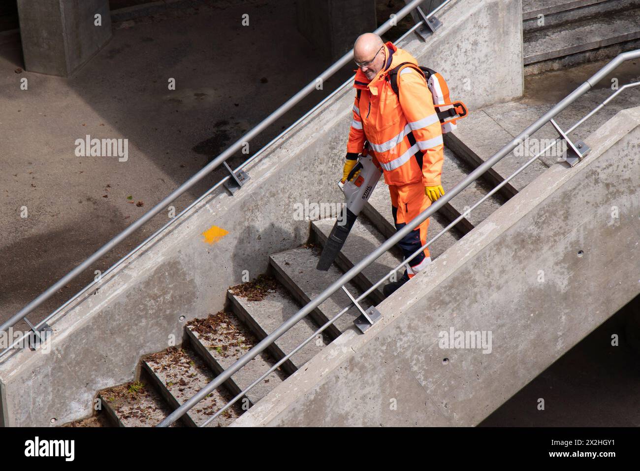 Basilea, Svizzera - 18 aprile 2024: Un lavoratore che indossa l'uniforme arancione per la pulizia delle scale del ponte cittadino con soffiatore Foto Stock