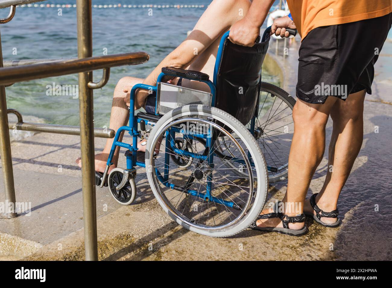 Assistente maschile che aiuta un uomo su una sedia a rotelle a godersi il mare su una rampa di accesso all'acqua. Spiaggia accessibile con sedia a rotelle. Foto Stock