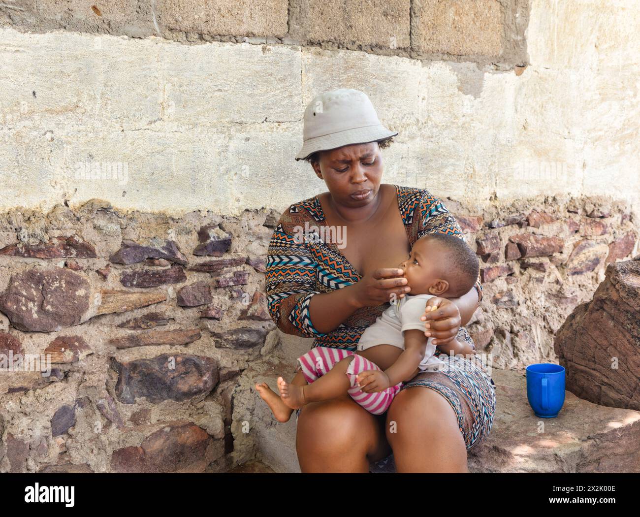 donna africana del villaggio che dà da mangiare al suo bambino all'aperto, seduta di fronte alla casa nel cortile Foto Stock