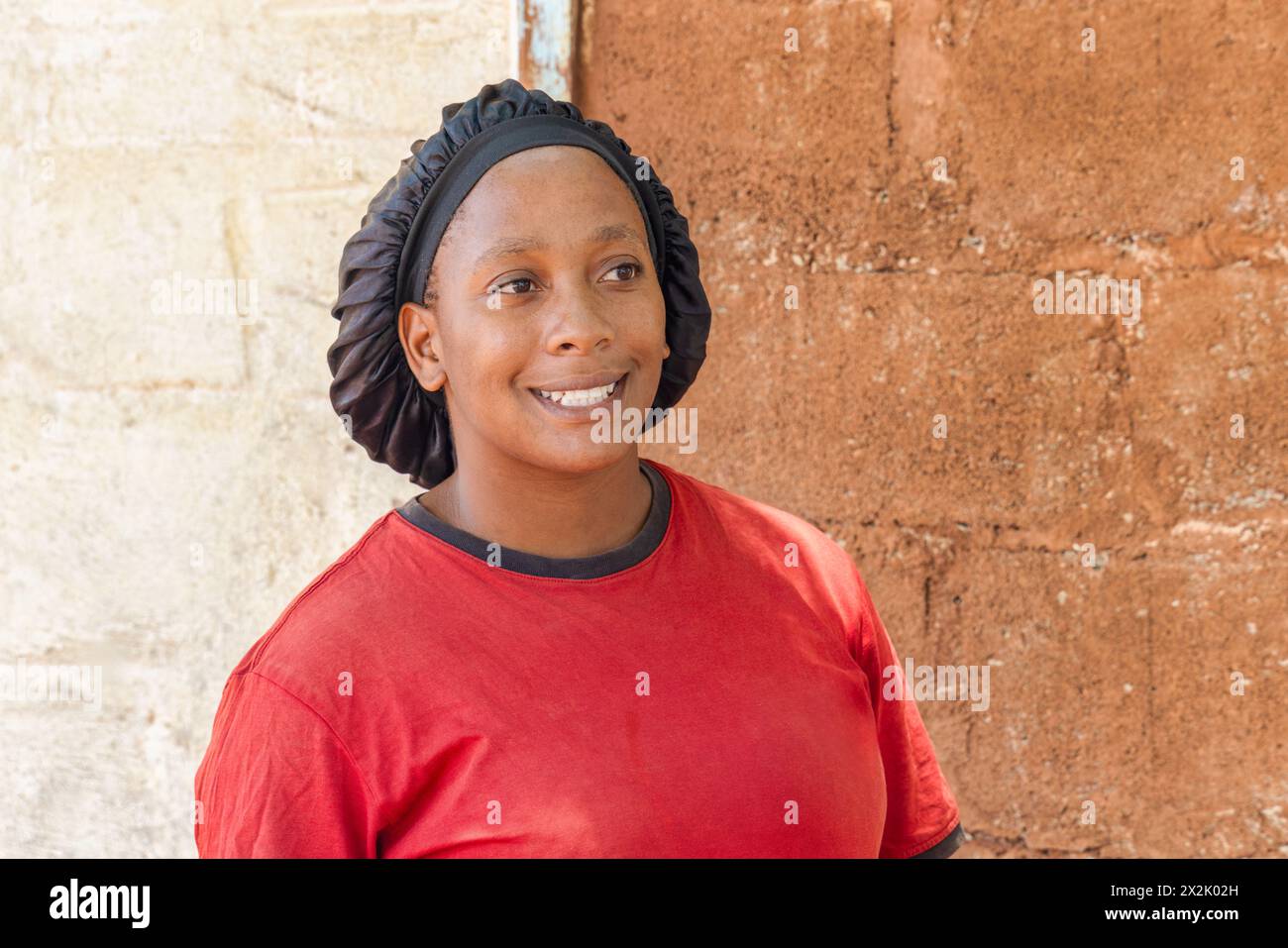 giovane donna africana sorridente con copricapo in piedi di fronte alla casa nella povera cittadina, insediamento informale Foto Stock