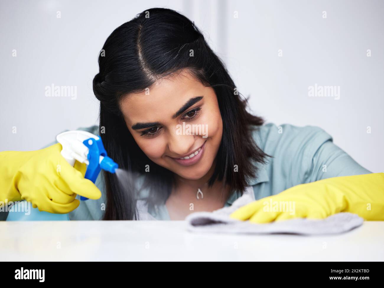 Donna indiana, sorriso o detergente con flacone spray per tavolo, bancone da cucina o mobili sporchi in casa. Felice, cameriera o inserviente che lava la superficie disordinata Foto Stock