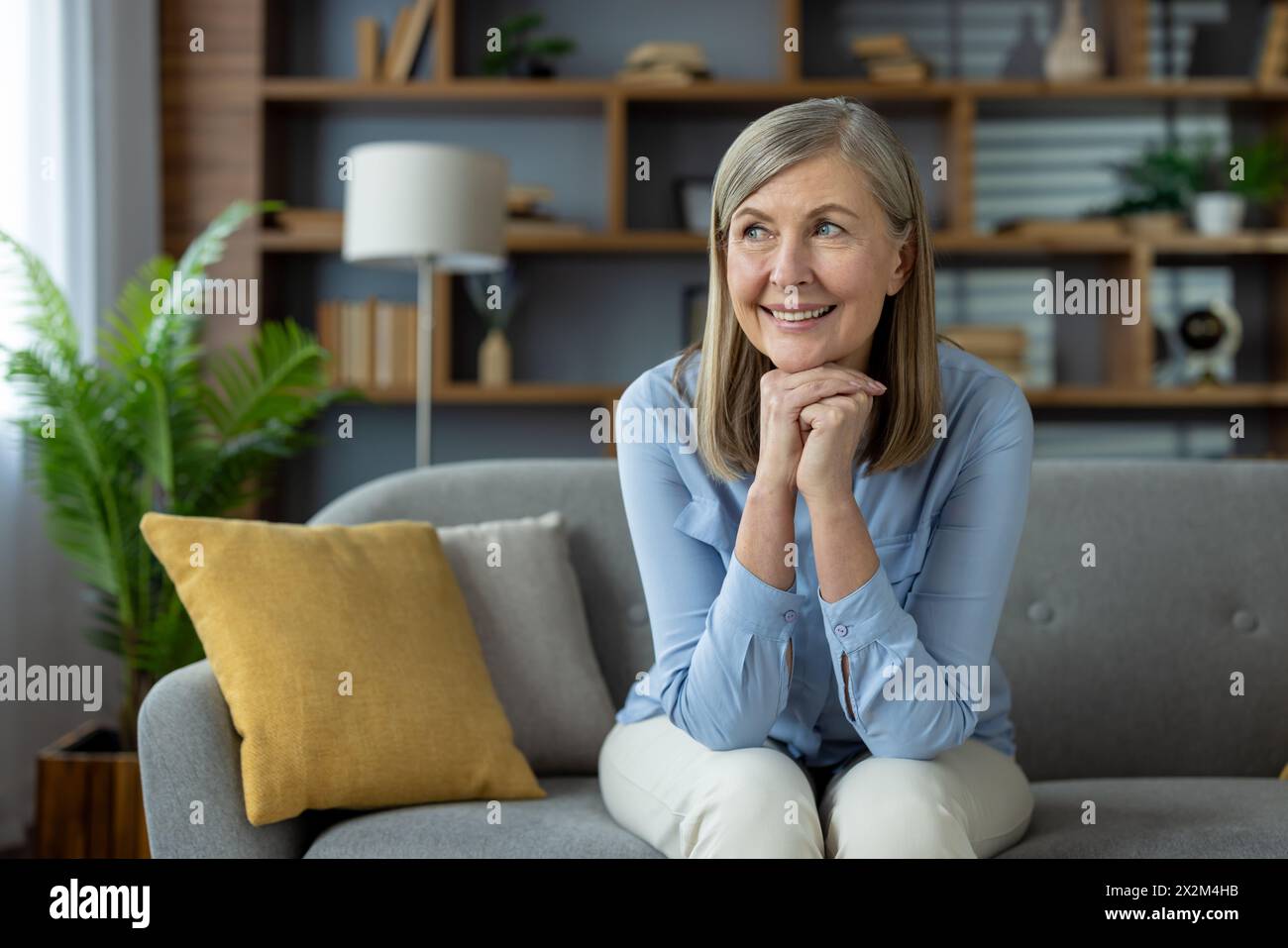 Una donna matura gode di un momento di pace a casa. Seduto comodamente su un divano, il suo volto sorridente esprime appagamento e gioia, circondato da un soggiorno arredato con gusto. Foto Stock