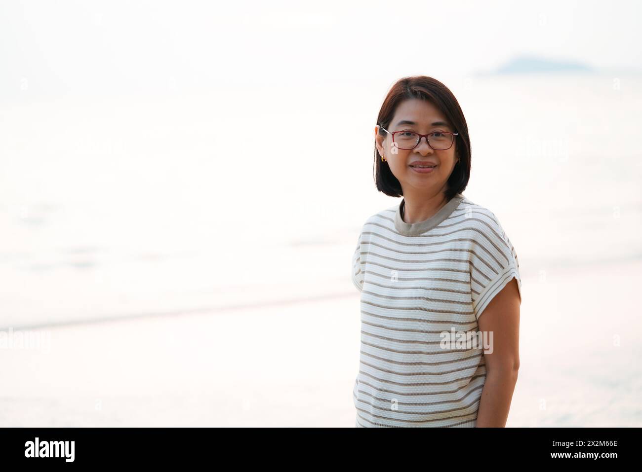 Ritratto di una sana donna asiatica di mezza età in piedi su una spiaggia, luce brillante da dietro, spazio per la copia e il design. Foto Stock