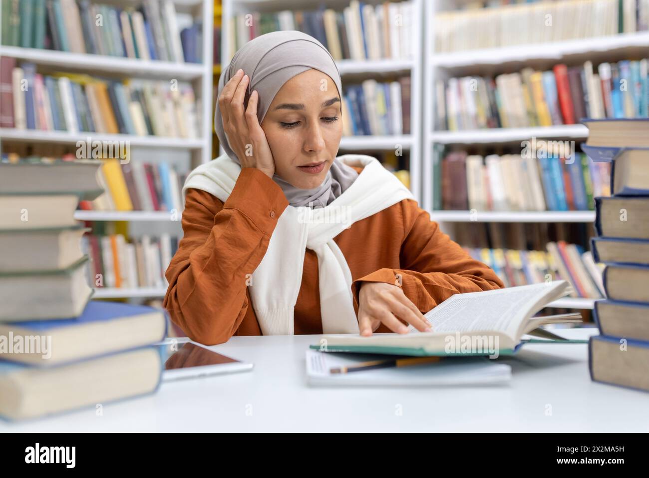 Una donna musulmana premurosa in un hijab immersa nei suoi studi in una biblioteca, circondata da mucchi di libri, che mostra dedizione e ricerca della conoscenza. Foto Stock