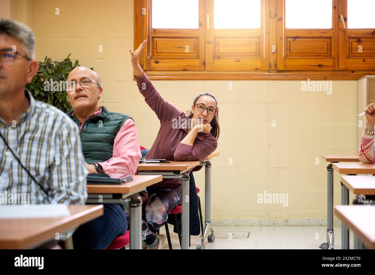 Una donna anziana alza con entusiasmo la mano in una classe tra colleghi, incarnando il concetto di apprendimento permanente e partecipazione attiva Foto Stock