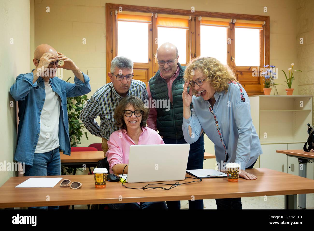 Un gruppo di allegri adulti anziani si riunisce intorno a un computer portatile con espressioni animate, condividendo un'esperienza di apprendimento in un accogliente ambiente scolastico Foto Stock