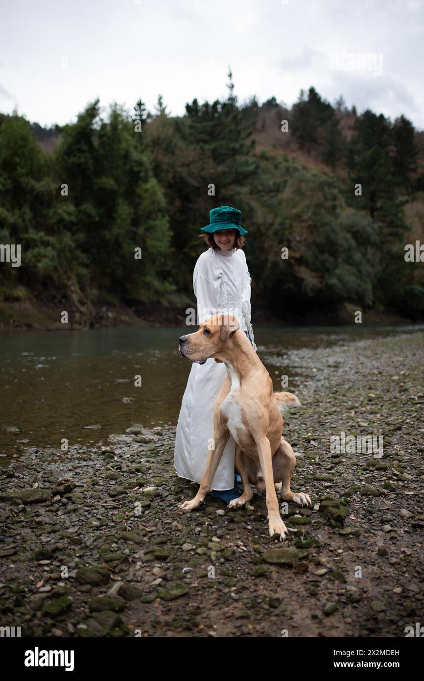Una donna con un abito bianco e un elegante cappello verde gode di un momento di pace con il suo fedele grande cane abbronzato in riva al fiume, circondato da alberi Foto Stock