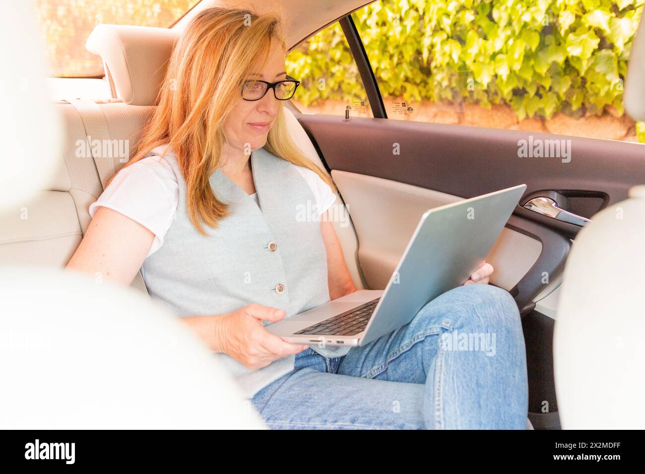 Una donna di mezza età lavora in remoto sul proprio laptop mentre è seduta comodamente sul sedile posteriore di un'auto, sottolineando la flessibilità del lavoro moderno Foto Stock