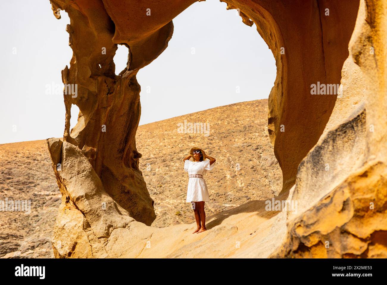 Una donna con un abito bianco e un cappello si erge pacificamente all'interno di un arco di roccia intemprato in un paesaggio deserto, incarnando tranquillità e relax. Foto Stock