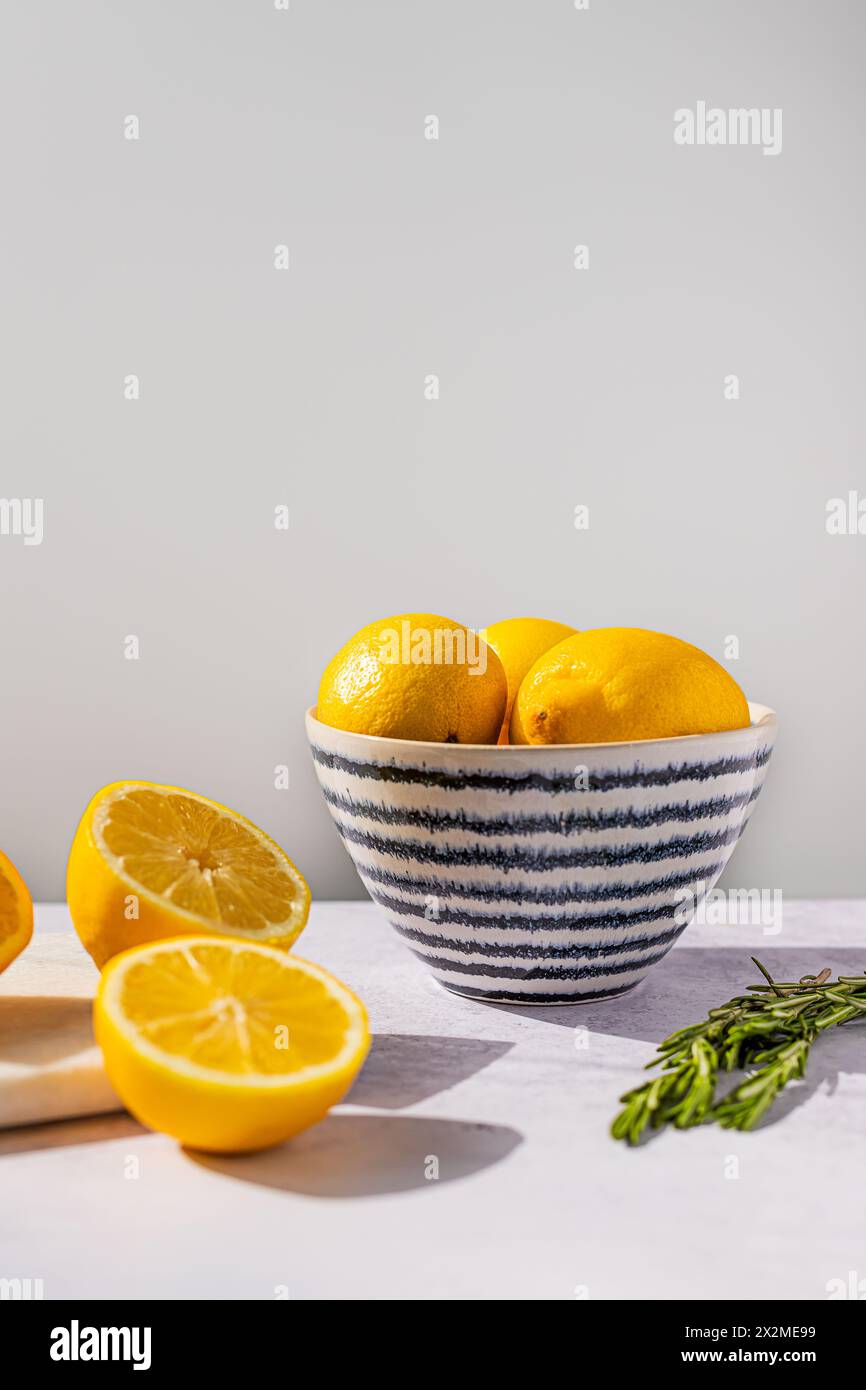 Una ciotola in ceramica con strisce verticali ripiene di limoni gialli luminosi, con frutta tagliata a metà ed erbe fresche su una superficie leggera Foto Stock