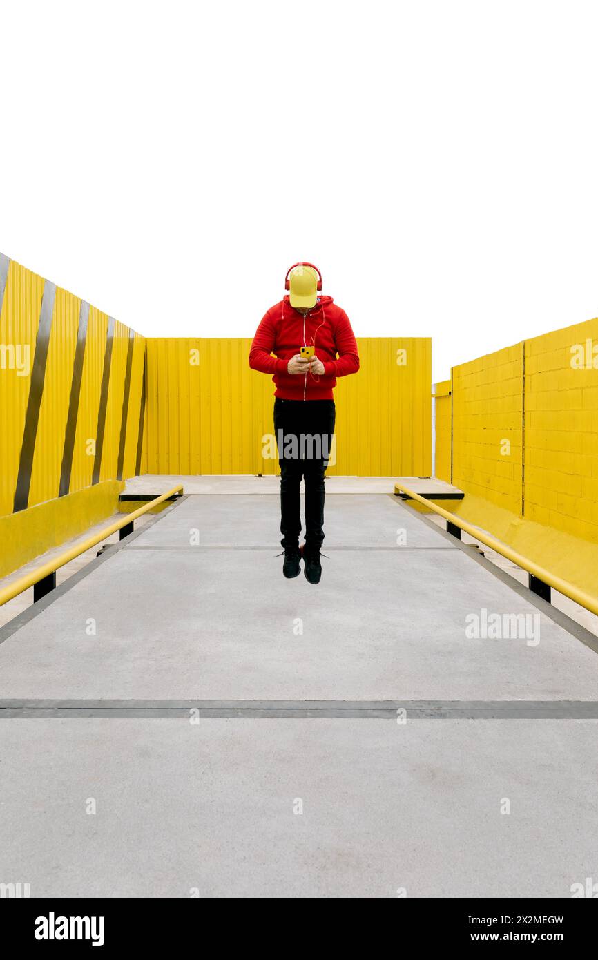 Un uomo con cappuccio rosso e jeans si trova su una passerella gialla, che utilizza uno smartphone e ascolta le cuffie Foto Stock