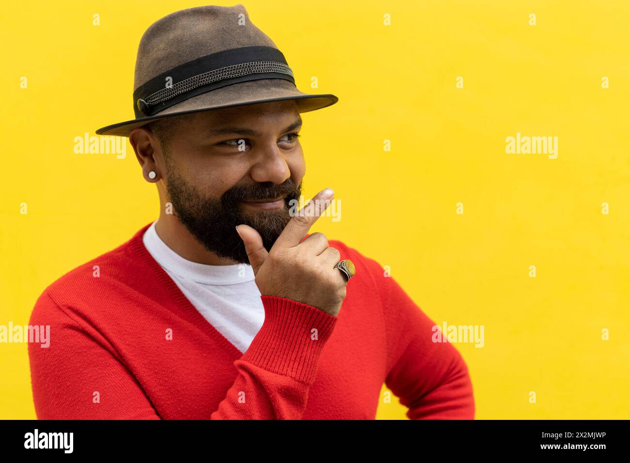 Uomo latino adulto (39) con barba, guarda alla sua destra con un gesto ottimista. Indossa cappello, maglione rosso e flanella bianca. Sfondo giallo Foto Stock
