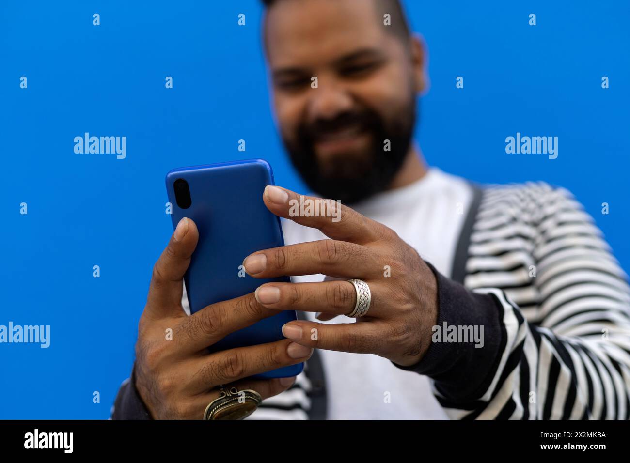 Maschio adulto (39) con barba, colpo medio corto, messa a fuoco sulle mani, uso del cellulare sorridente. Abito in maglione a righe, t-shirt bianca, wal blu Foto Stock