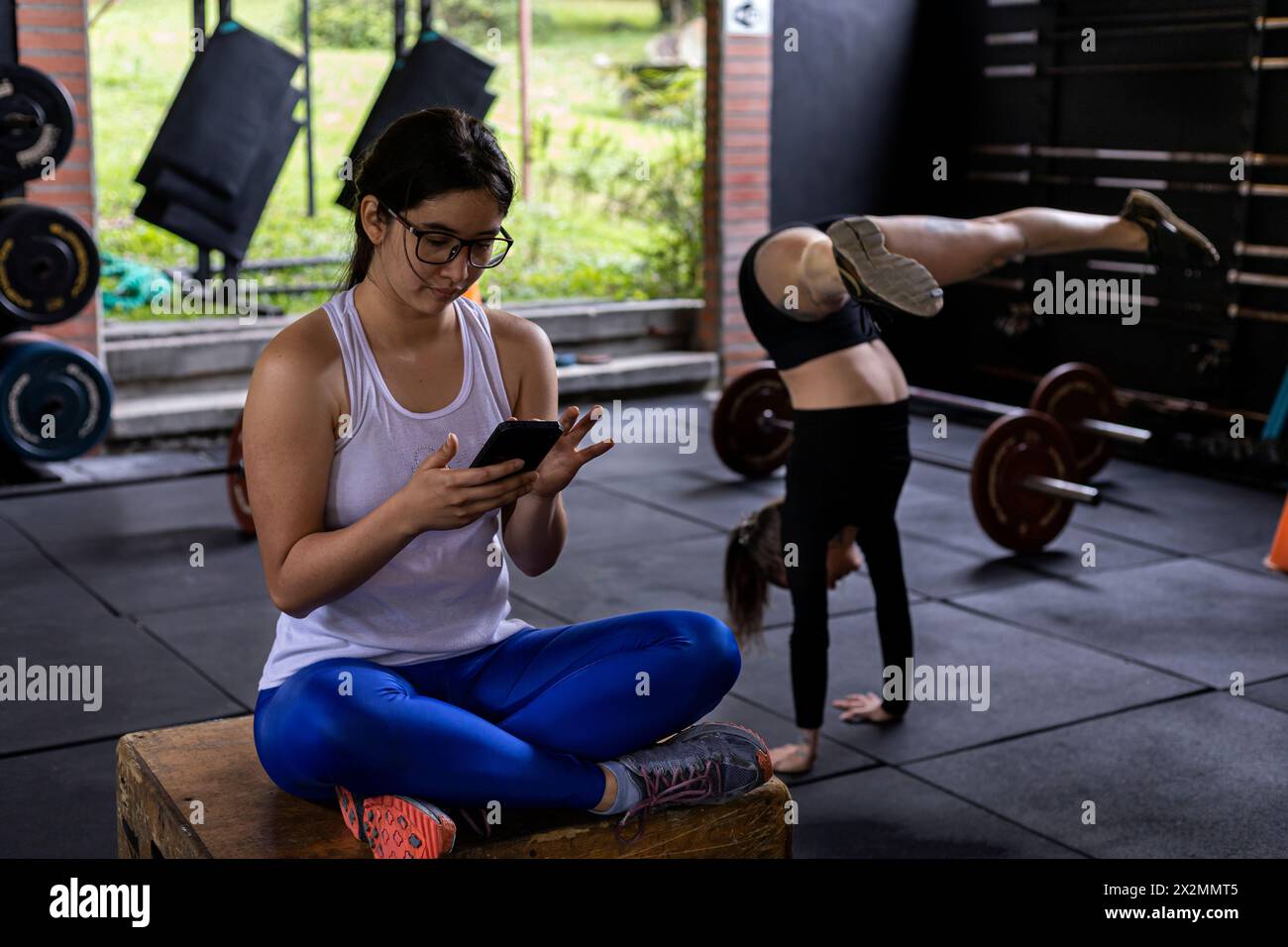 Giovane donna latinoamericana seduta con il cellulare in mano a controllare i suoi messaggi mentre si riposa dal suo allenamento in palestra, accanto a lei un te Foto Stock