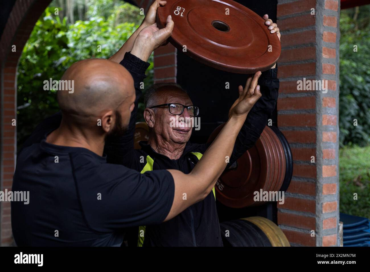 Un vecchio latinoamericano e il suo allenatore tengono in mano un disco di peso che usano per esercizi di riabilitazione e terapia in palestra. Healt Foto Stock