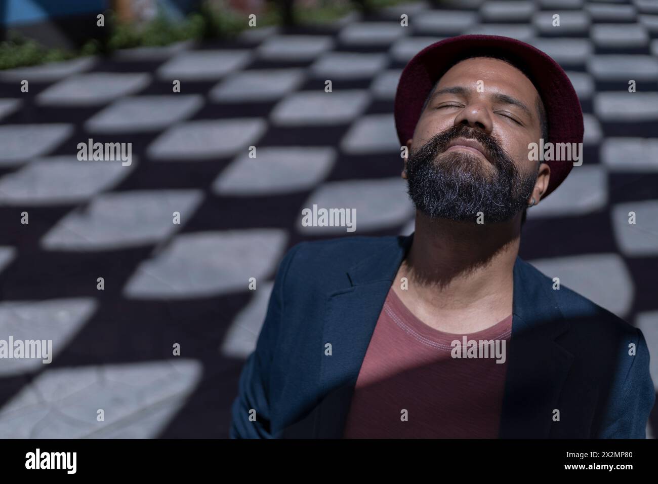 Ritratto dell'uomo bohémien latinoamericano con occhi chiusi, barba, cappello e giacca. Sfondo di ombre con forme rombo. Concetto di persone reali. Foto Stock