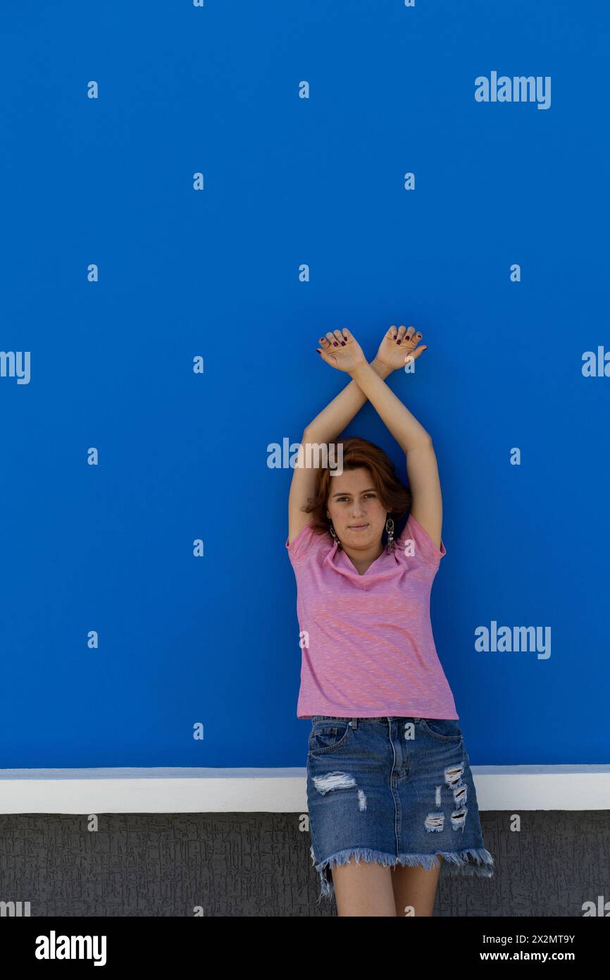Scatto medio lungo di una giovane donna (22) rossa dell'America Latina che guarda e guarda la fotocamera con le braccia rivolte verso l'alto. Sfondo blu della parete. Verticale c Foto Stock