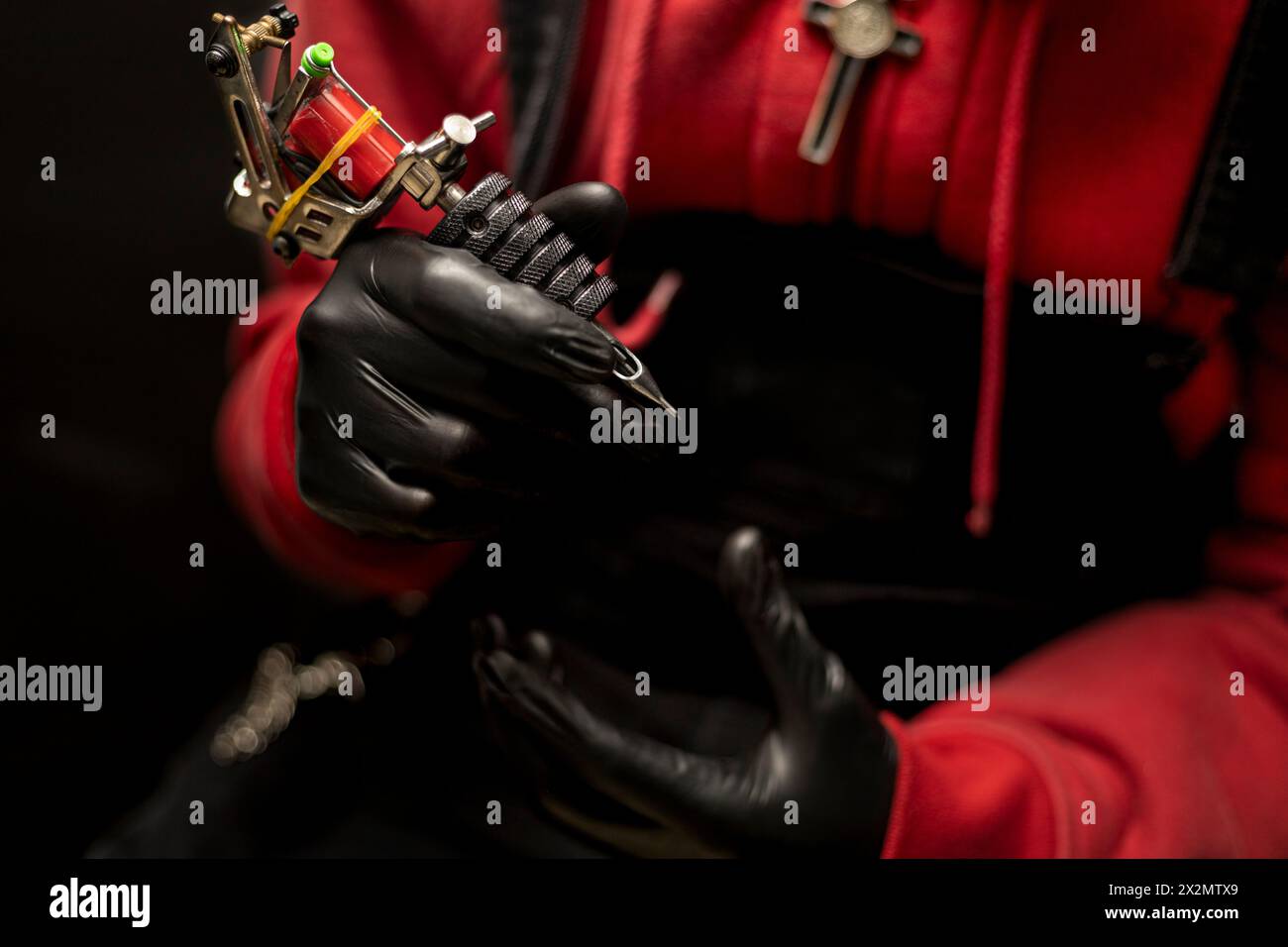 Mani di tatuatore che mostrano la sua macchina per tatuaggi. Indossa guanti neri, maglione rosso e sfondo nero. Concetto di Body art Foto Stock