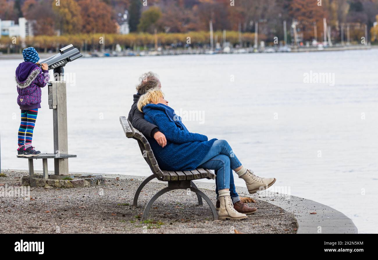 Ein Mann und eine Frau sitzen auf einer Parkbank beisamen und schauen auf den Bodensee. Daneben ein Kind, welches mit einem Fernglas Spielt. (Costanza Foto Stock