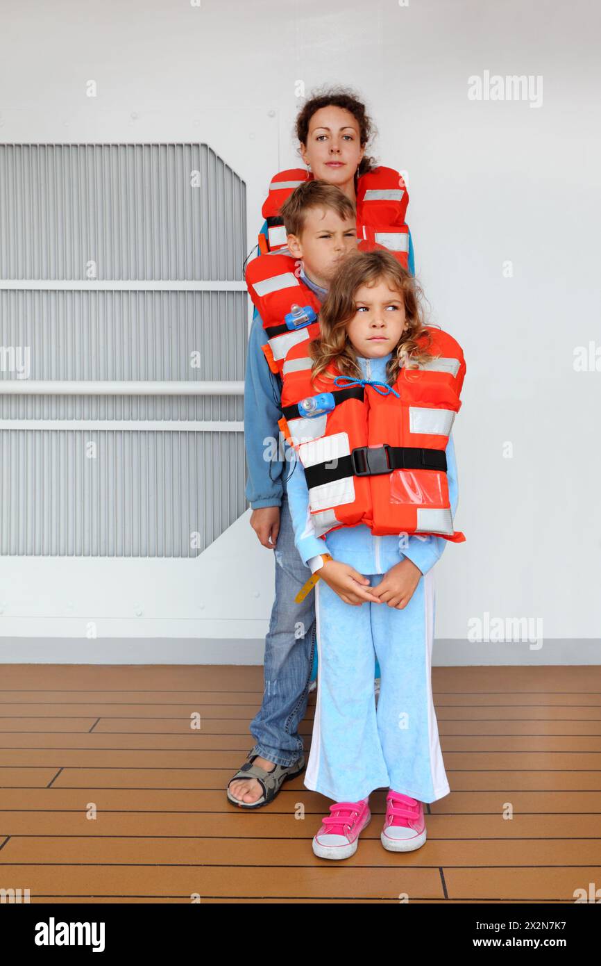 Madre, figlio e figlia testano i loro giubbotti di salvataggio arancioni sul ponte della nave da crociera; i bambini stanno di fronte alla mamma Foto Stock