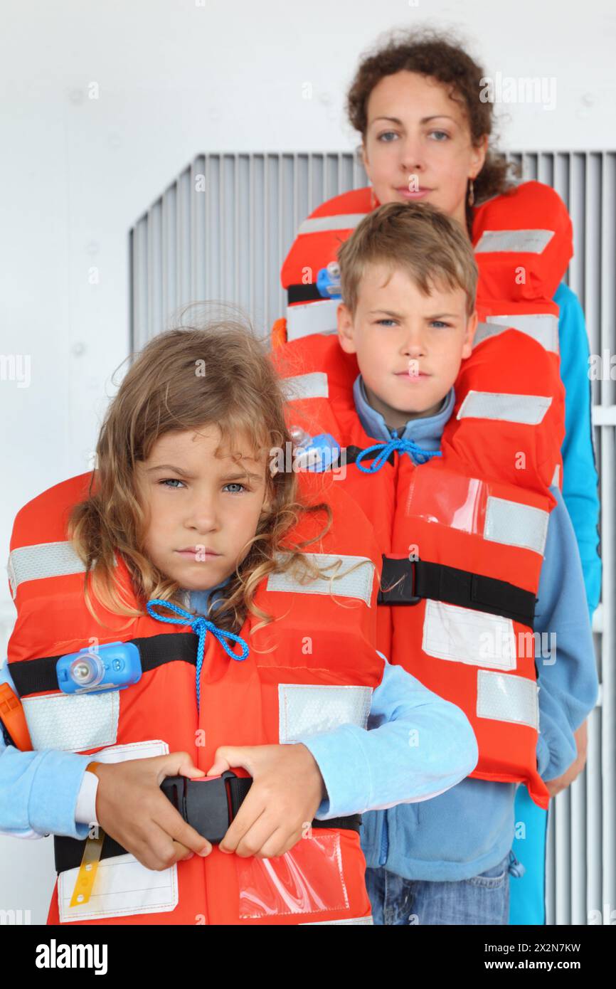 Madre, figlio e figlia testano i loro giubbotti di salvataggio arancioni sul ponte della nave da crociera; si concentrano sulla ragazza; i bambini stanno di fronte alla mamma Foto Stock