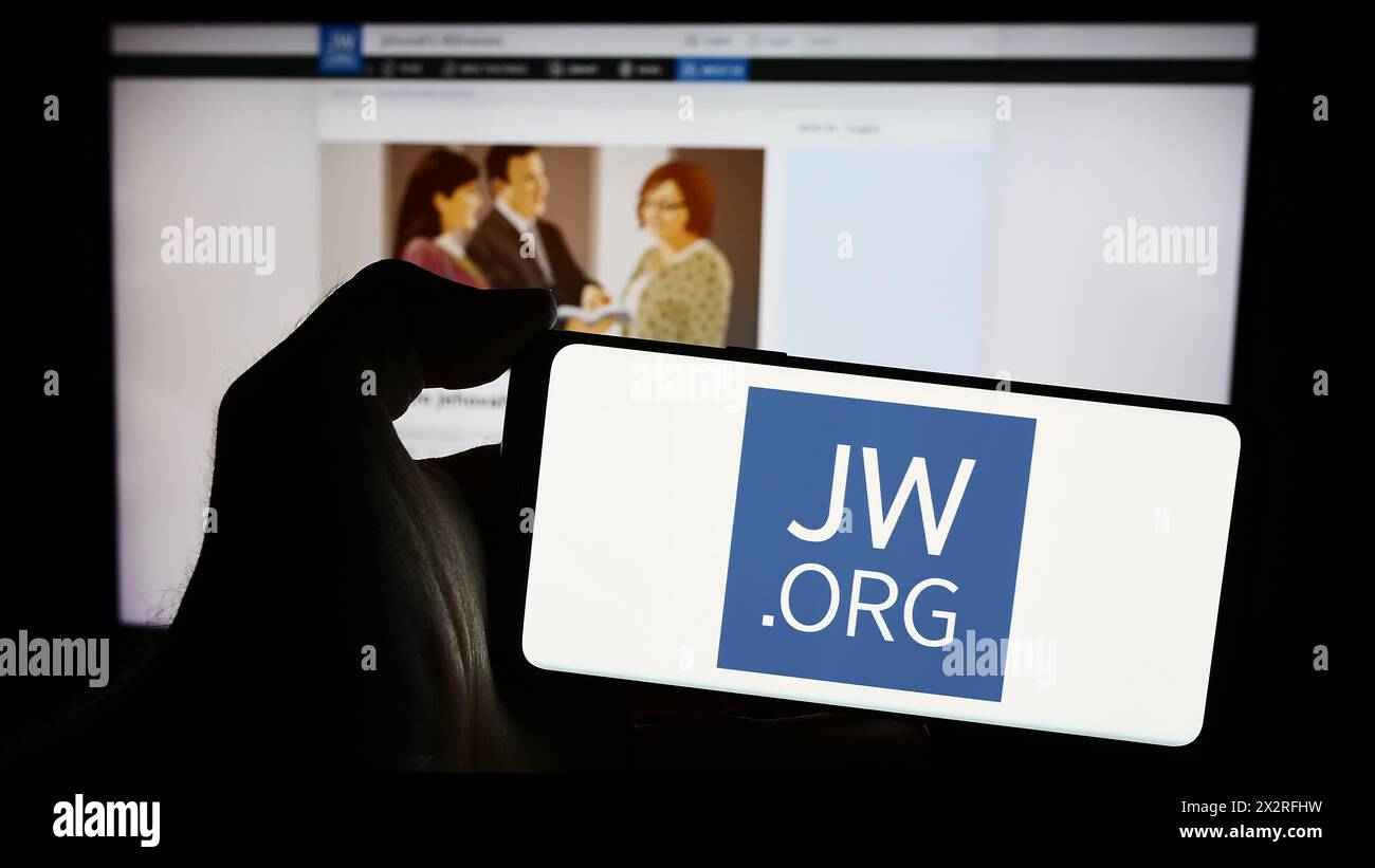 Persona che ha un cellulare con il logo dei testimoni dell'organizzazione religiosa di Geova davanti alla pagina web. Mettere a fuoco il display del telefono. Foto Stock
