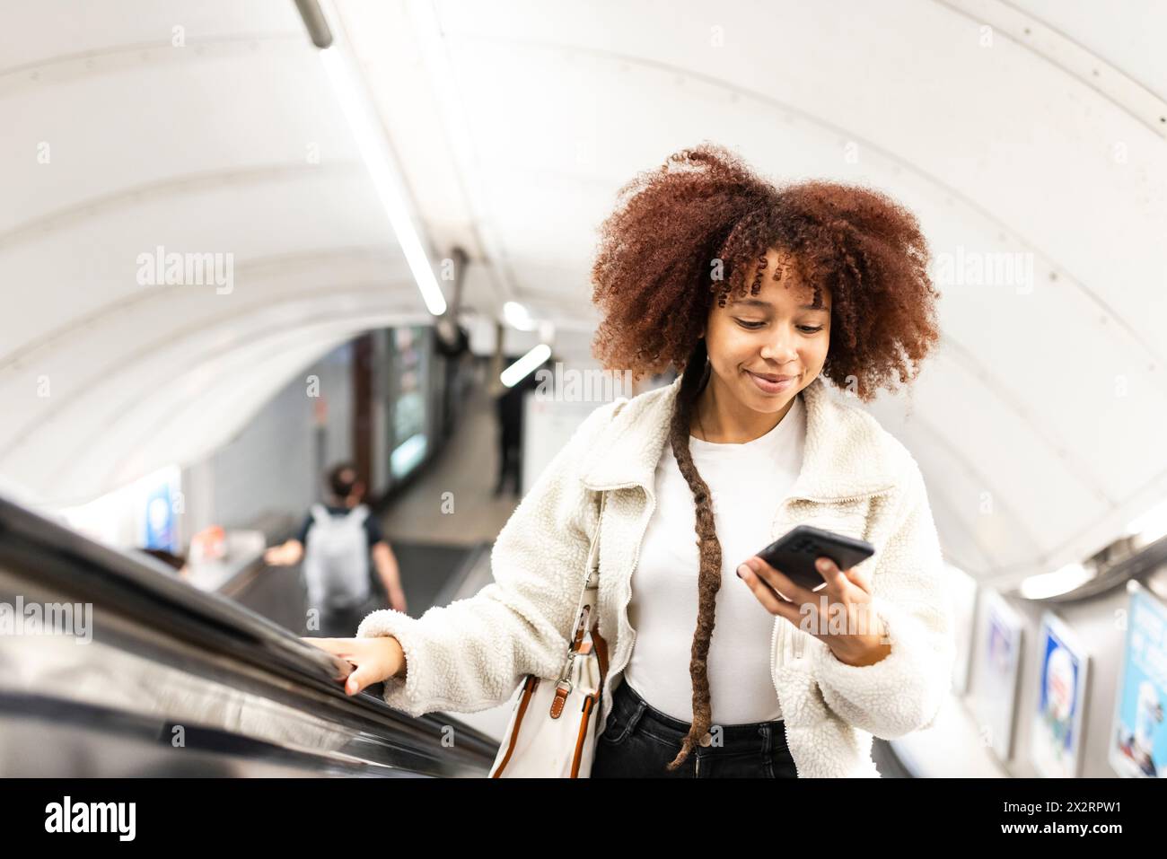 Donna sorridente che usa il cellulare sulla scala mobile della metropolitana Foto Stock