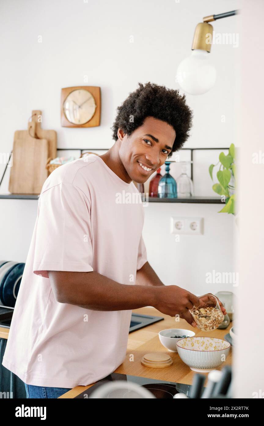 Uomo sorridente che prepara il muesli della colazione nel recipiente del bancone della cucina Foto Stock