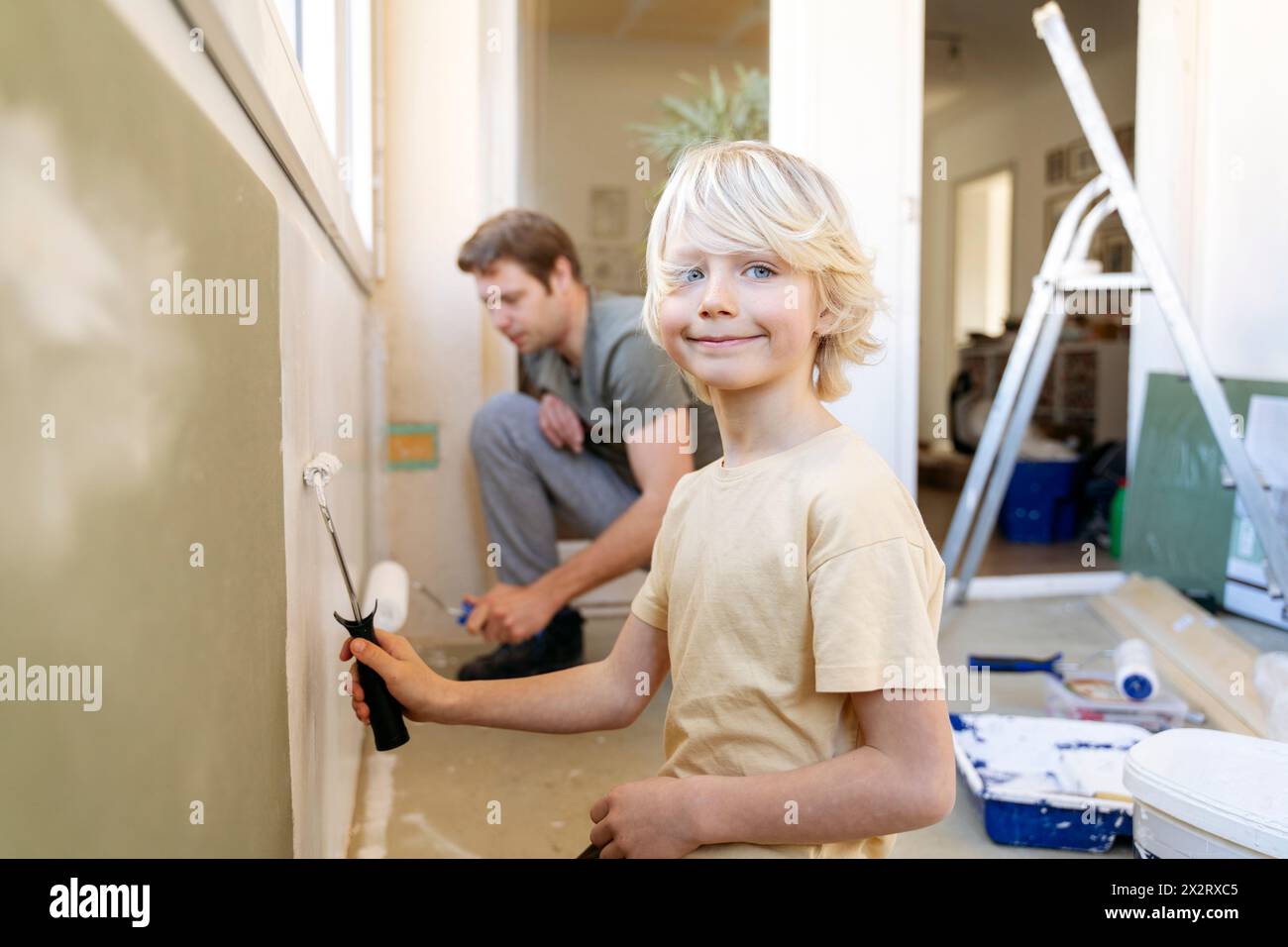 Un ragazzo sorridente che dipinge il muro con un rullo vicino al padre a casa Foto Stock