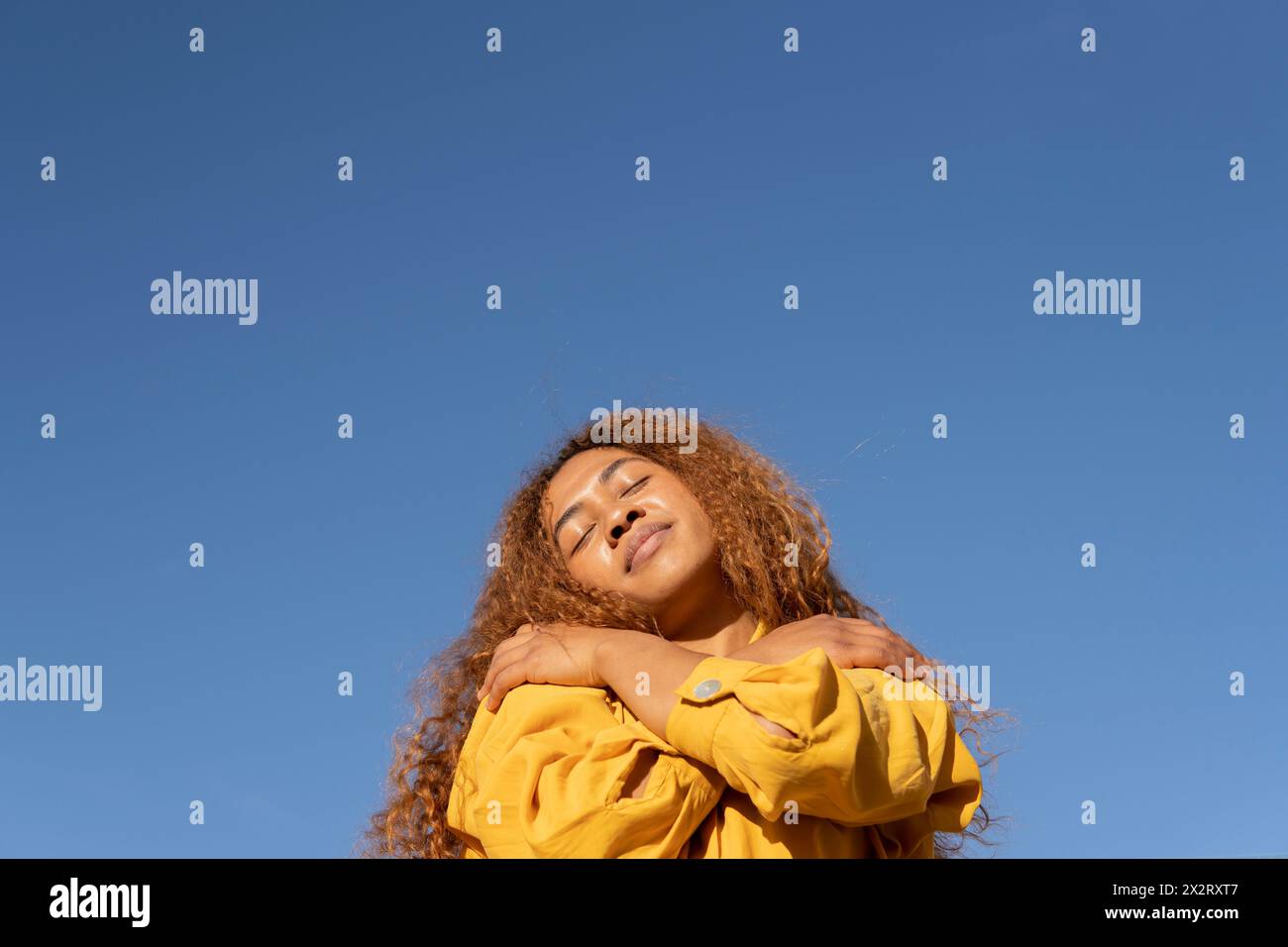 Giovane donna che si abbraccia con gli occhi chiusi sotto il cielo azzurro Foto Stock