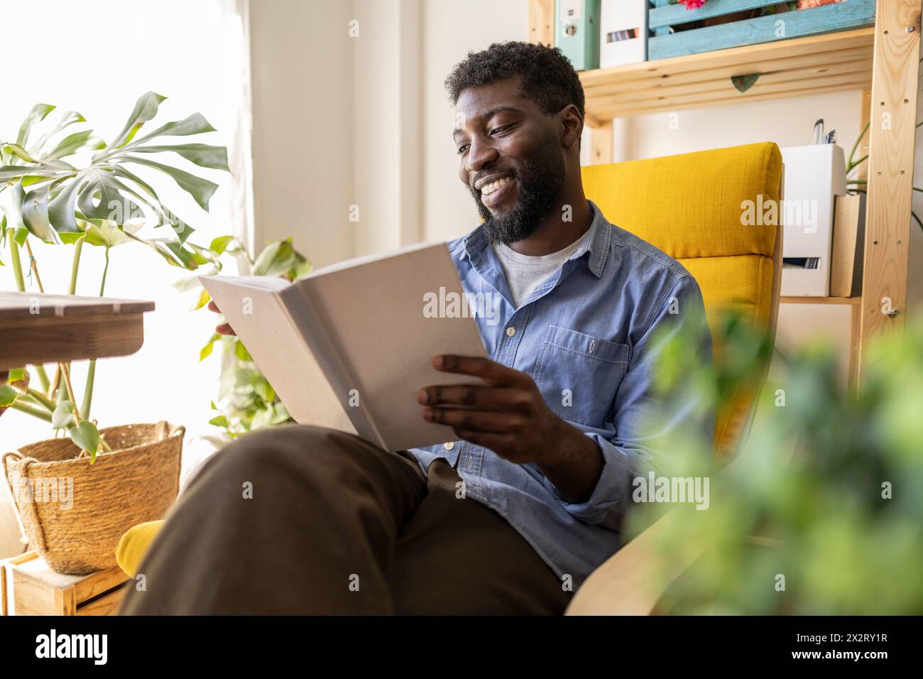 Felice persona non binaria che legge un libro sulla poltrona a casa Foto Stock