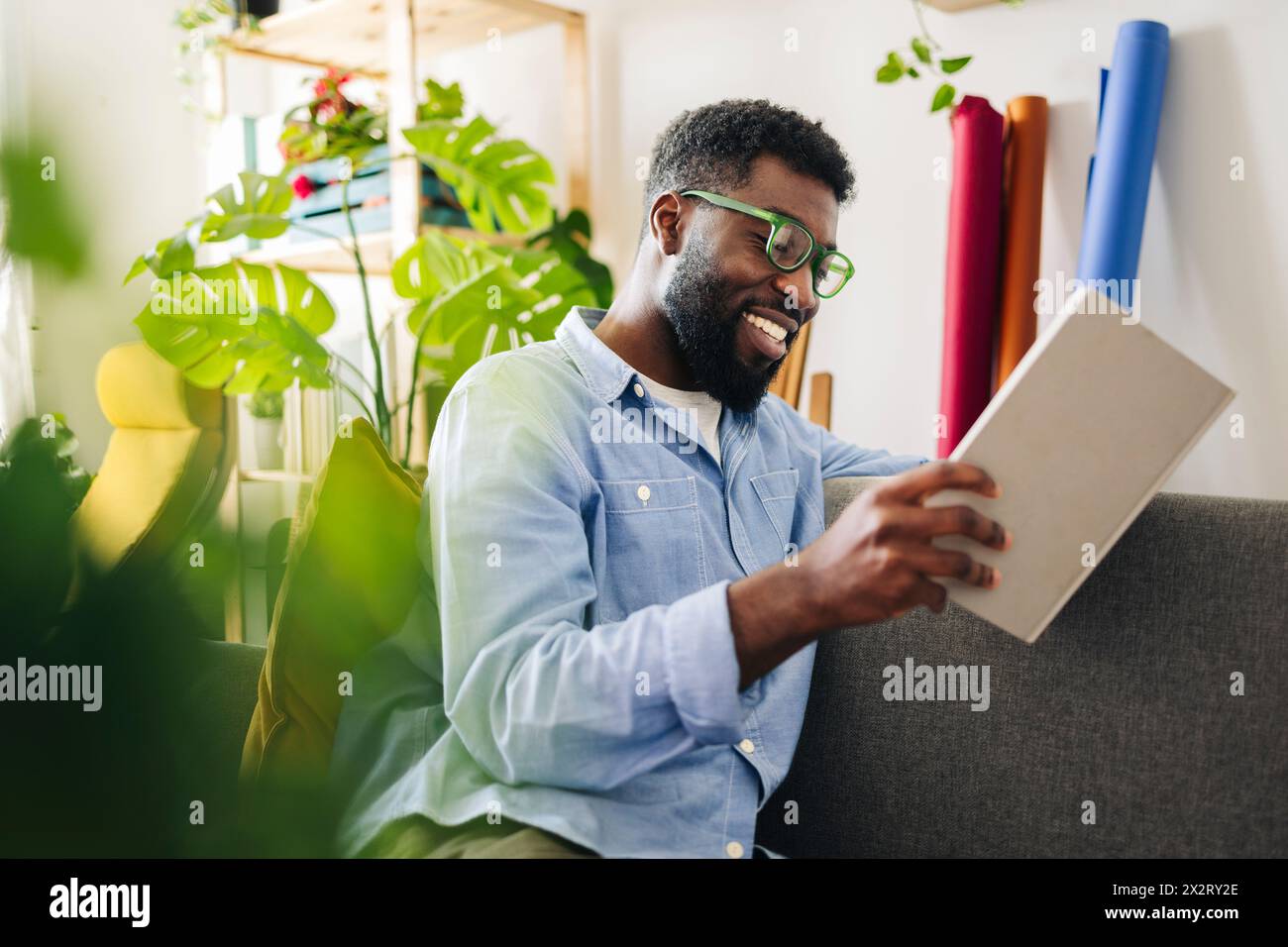 Felice persona non binaria che legge un libro nel soggiorno di casa Foto Stock