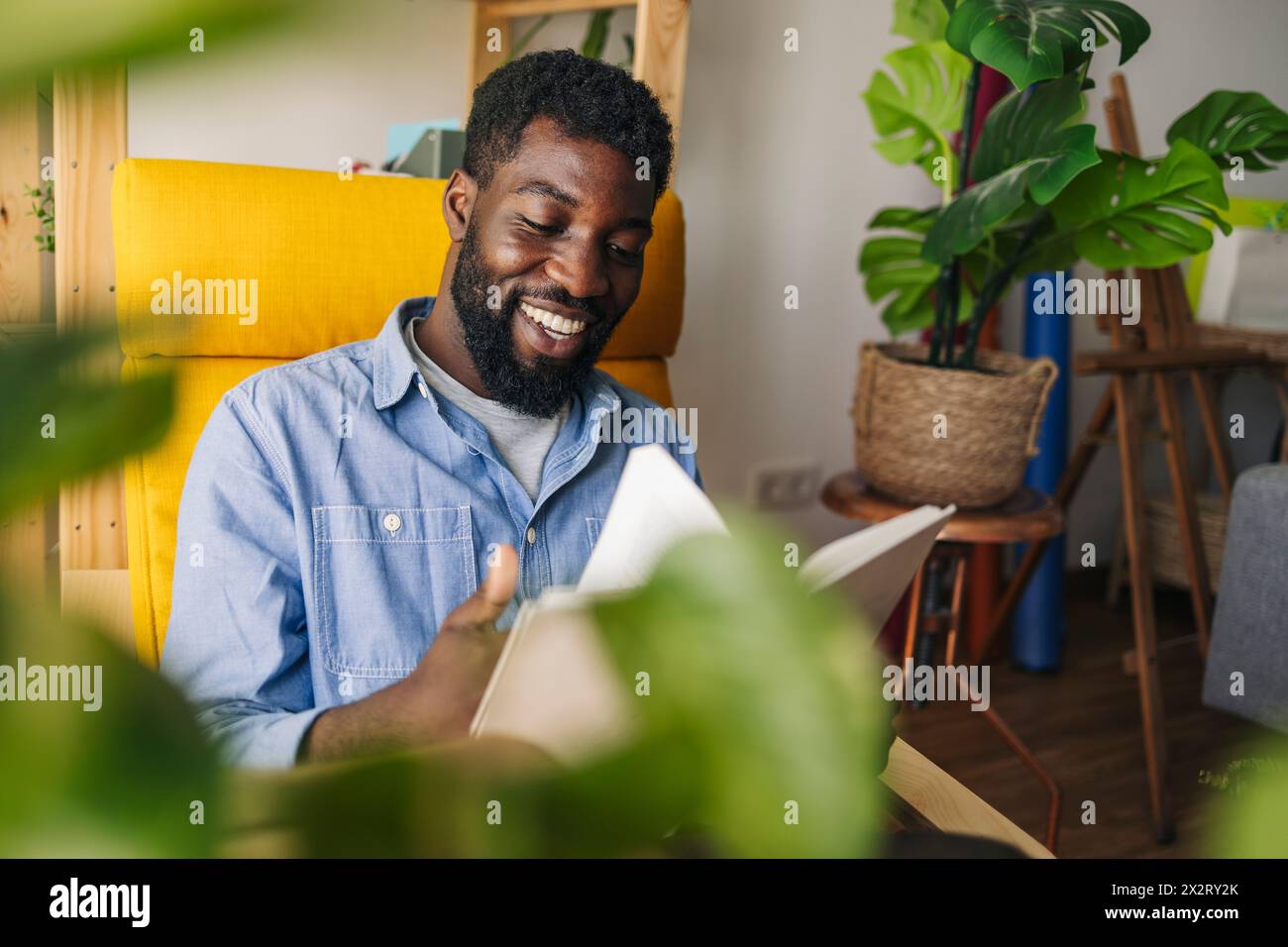 Felice persona non binaria che legge un libro vicino alle piante a casa Foto Stock