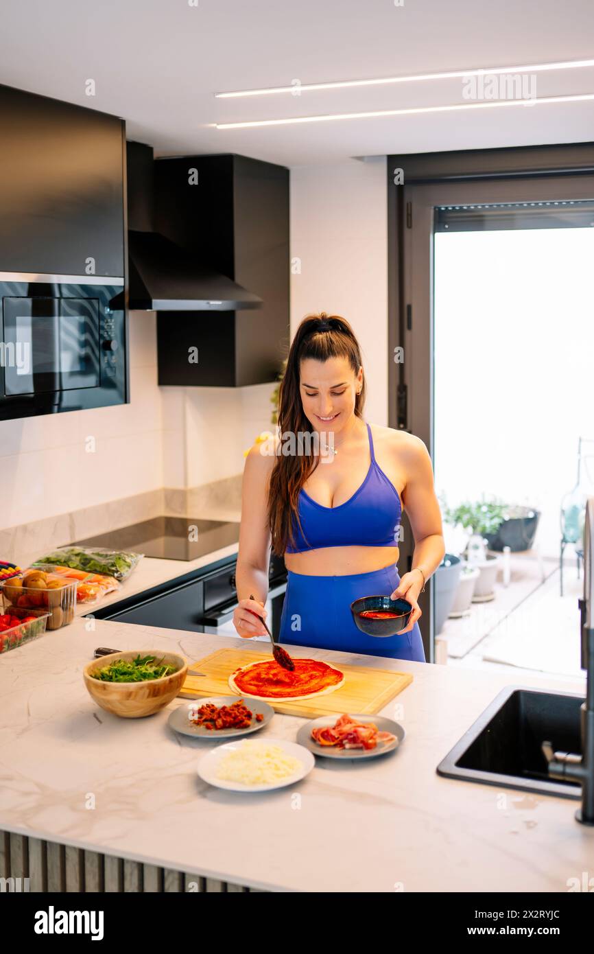 Donna sorridente che applica salsa rossa sulla base della pizza in cucina a casa Foto Stock